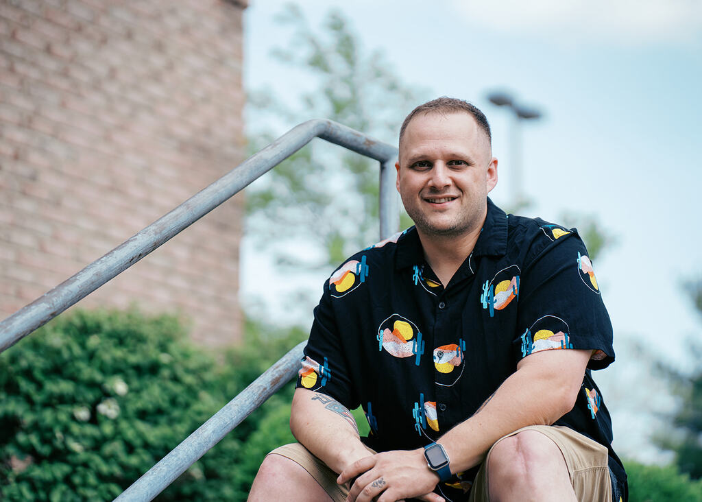 White male sitting on the top of stairs wearing a black shirt with catci and sunsets on it